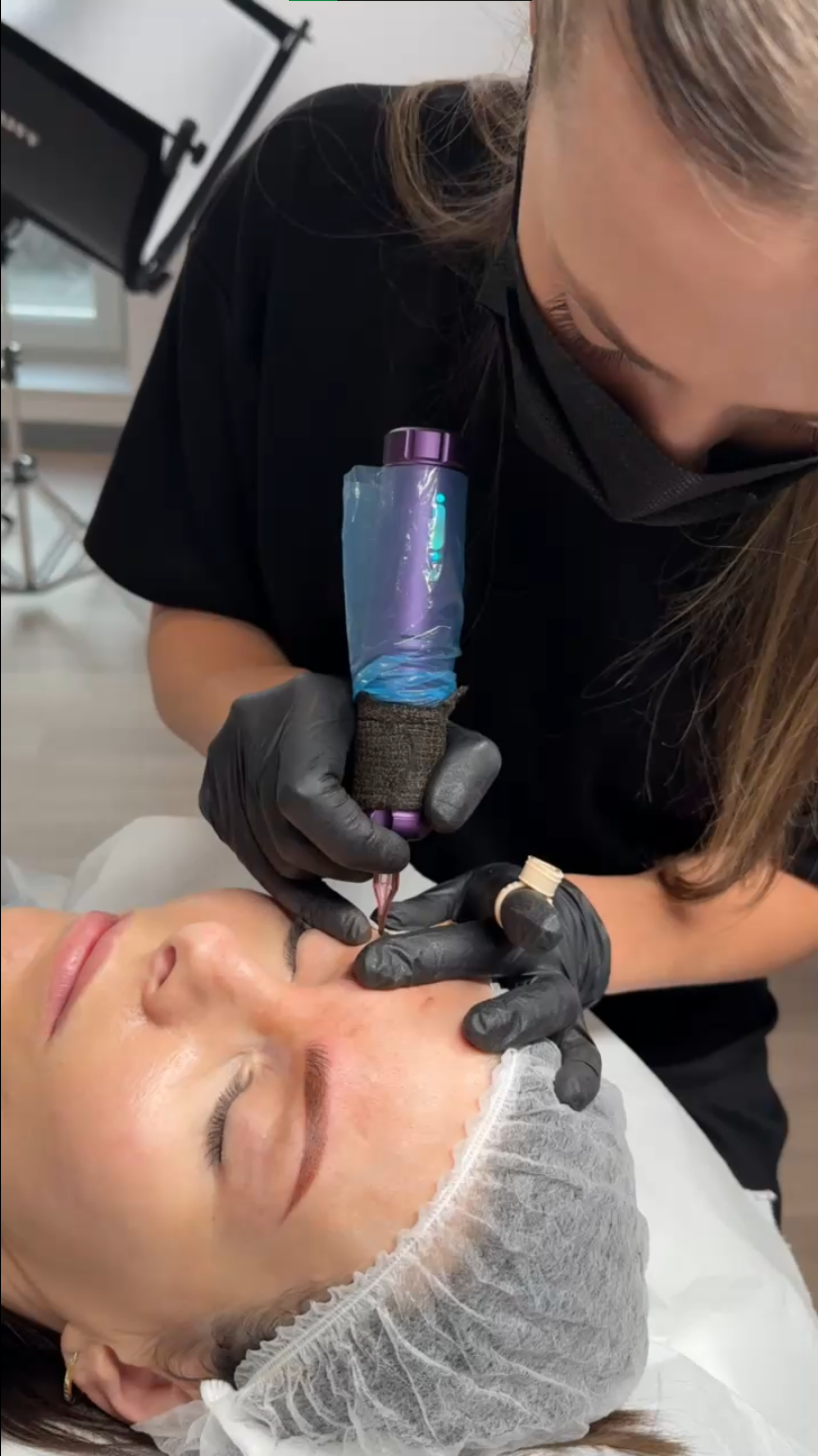 Kosmetyczka w czarnych rękawiczkach wykonuje makijaż permanentny brwi klientce leżącej na fotelu zabiegowym w czepku ochronnym.