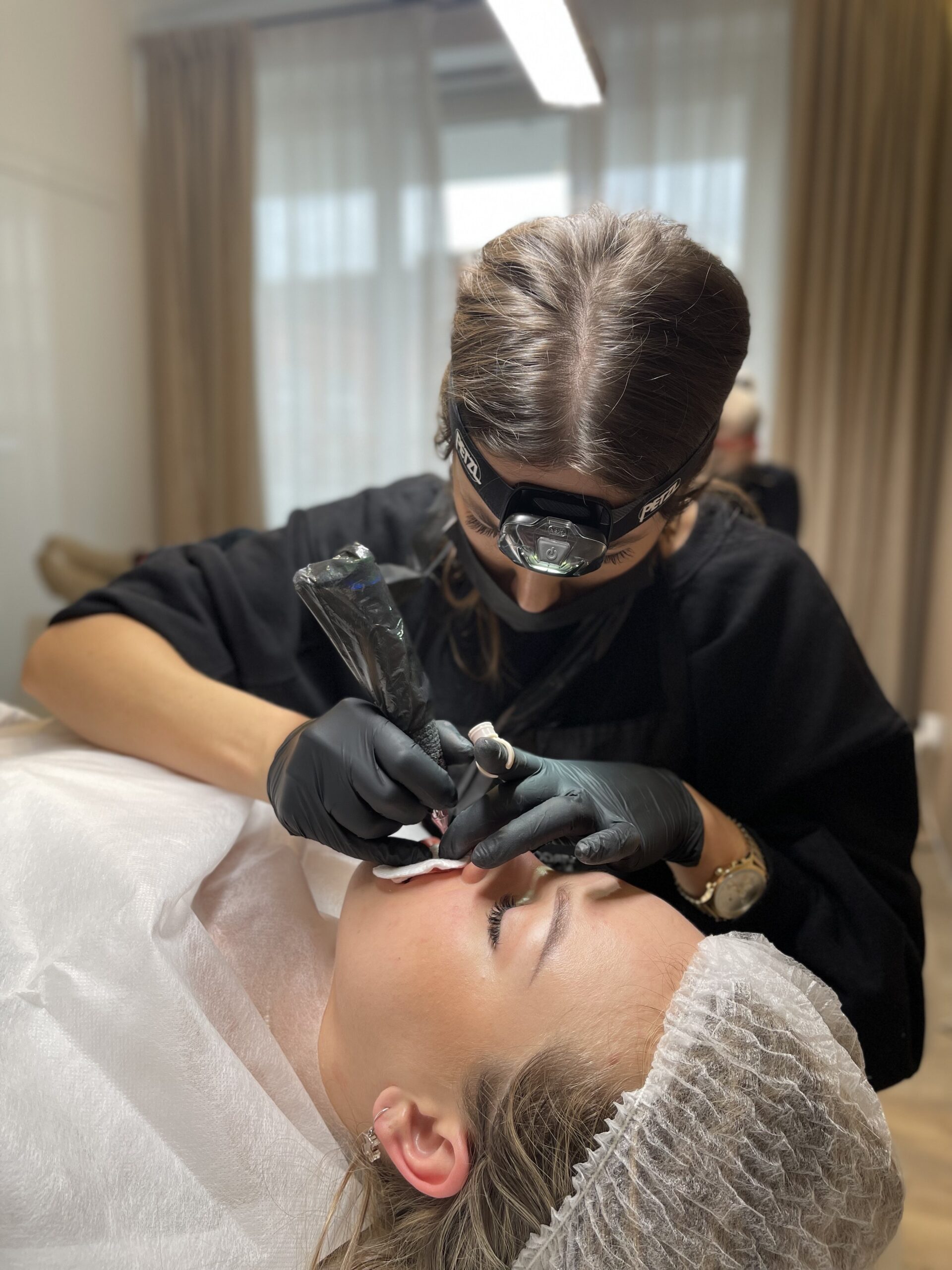 Kosmetyczka w rękawiczkach i lampą czołową wykonuje makijaż permanentny ust klientce leżącej w jednorazowym stroju ochronnym.