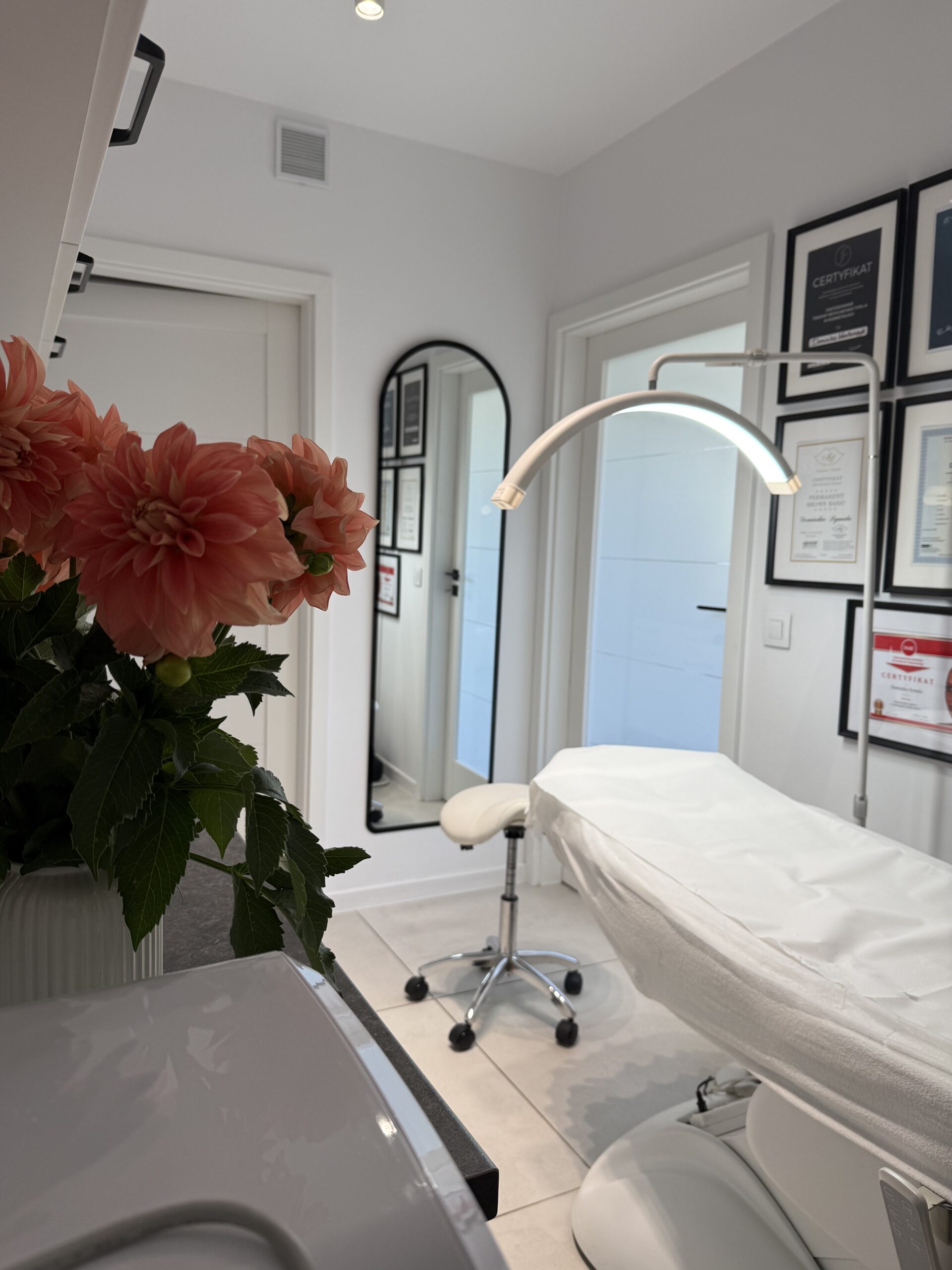 Nowoczesny gabinet kosmetyczny z fotelem zabiegowym, lampą i certyfikatami na ścianie, ozdobiony bukietem kwiatów.