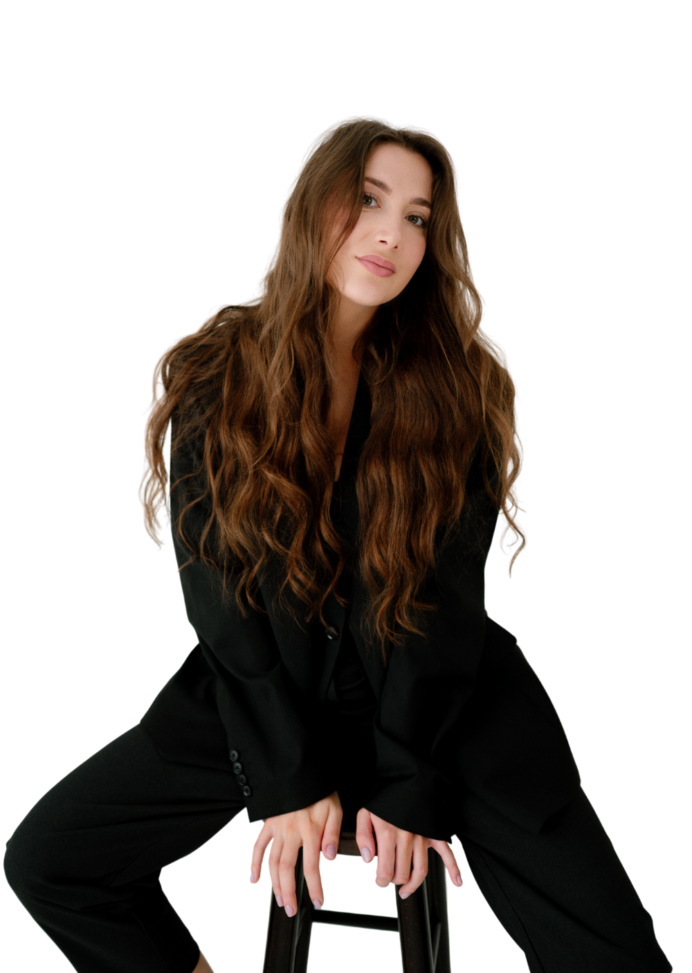 Kobieta z długimi, falowanymi włosami w czarnym garniturze siedząca na stołku na tle ściany.
